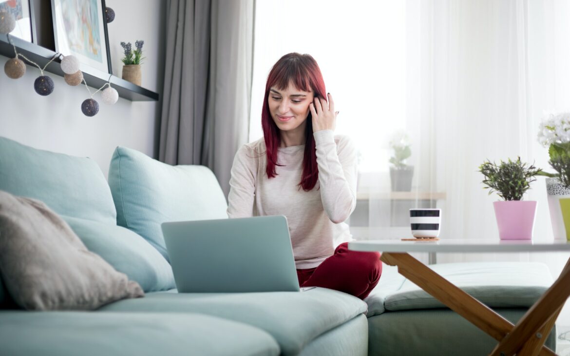 Cheerful woman browsing internet with laptop while sitting on sofa at home